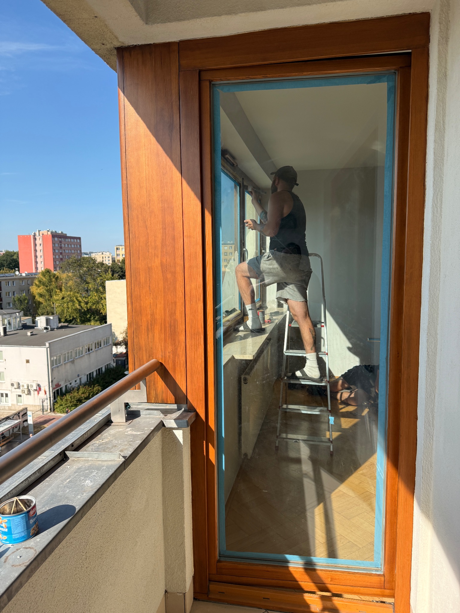 Mężczyzna na drabinie uszczelnia okno balkonowe z drewnianą ramą, z widokiem na miasto. Robocze akcesoria na parapecie.