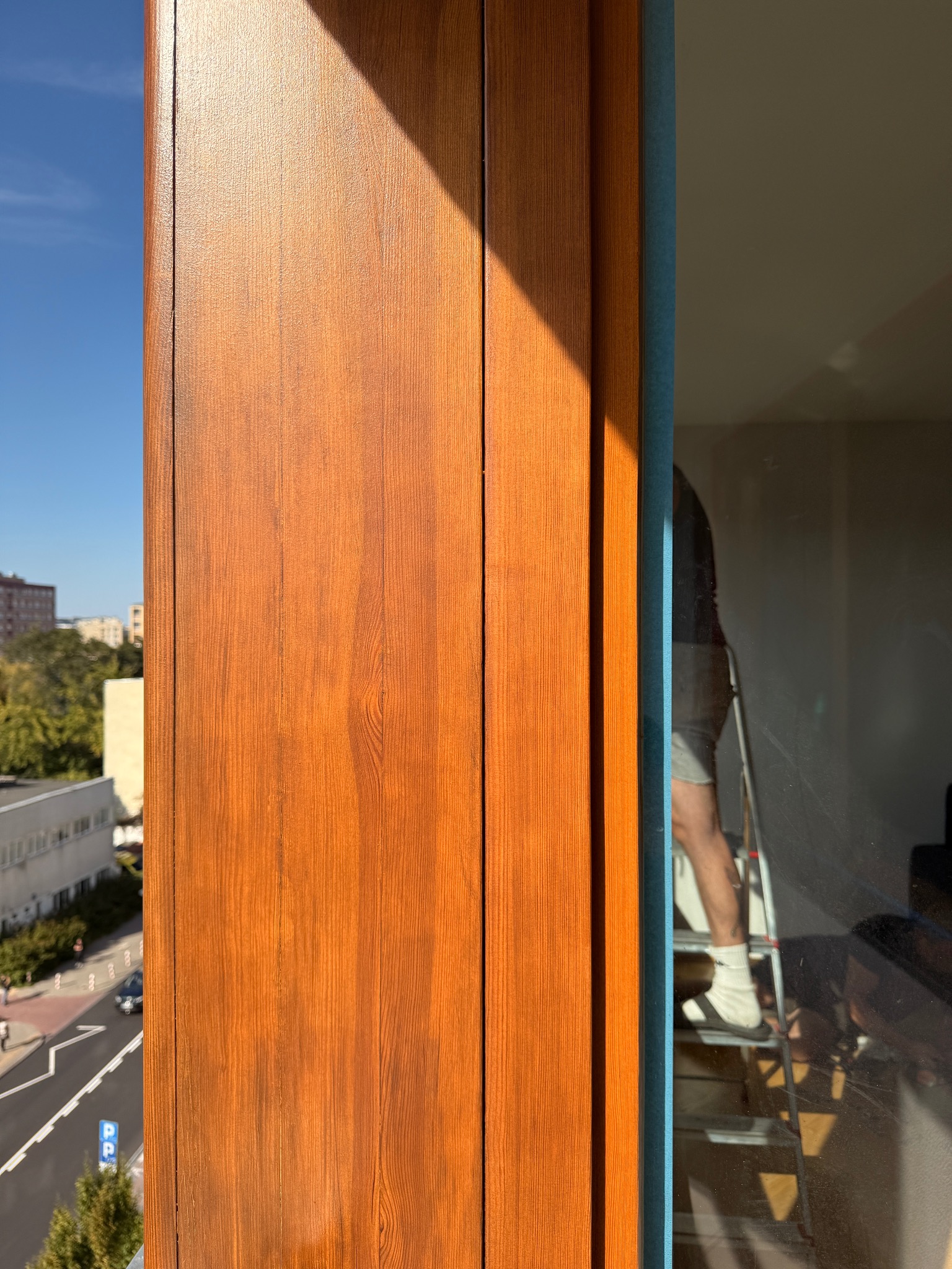 Detal wykończenia okna z drewnianą okleiną w ciepłym odcieniu brązu, widok z balkonu na ulicę i budynki, odbicie osoby na drabinie w szybie okiennej.