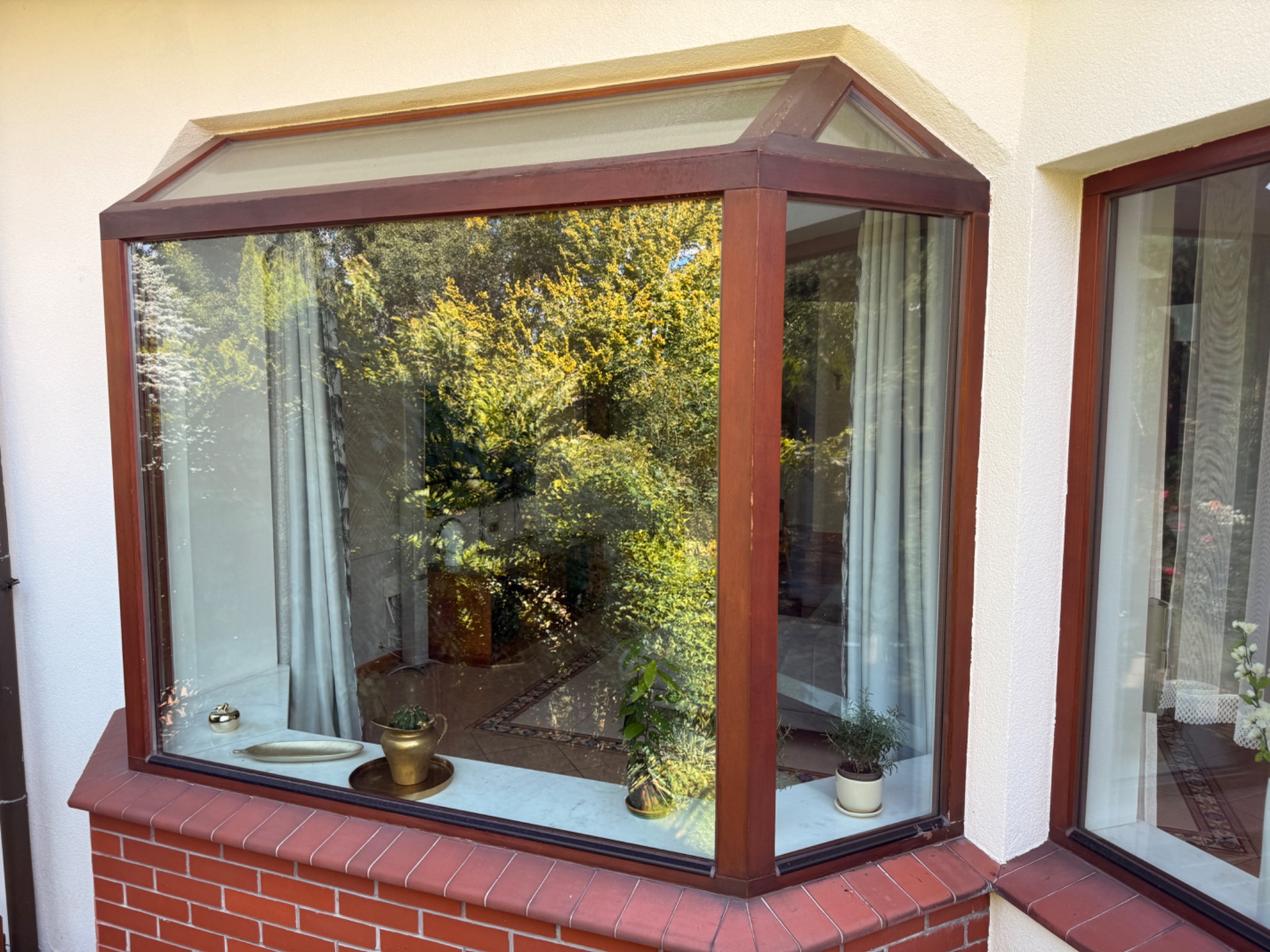 Kątowe okno z brązowymi ramami, widok na zieleń z zewnątrz, parapet z cegły i rośliny doniczkowe. Widoczne firanki w oknach.