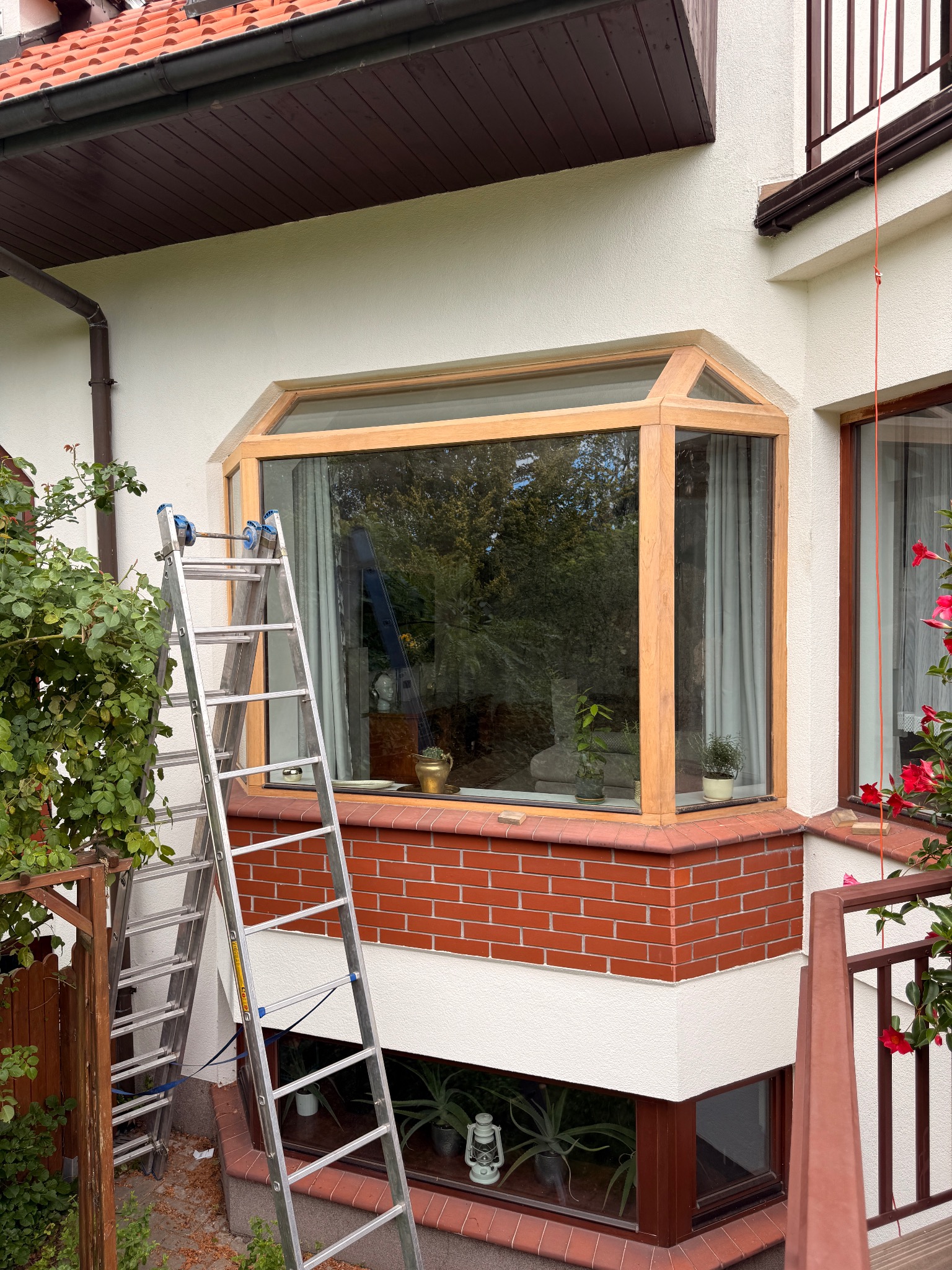 Okna wykuszowe z drewnianą ramą, widok z zewnątrz. Przystawiona drabina sugeruje prace naprawcze lub konserwacyjne. Ceglana podmurówka i roślinność w tle.