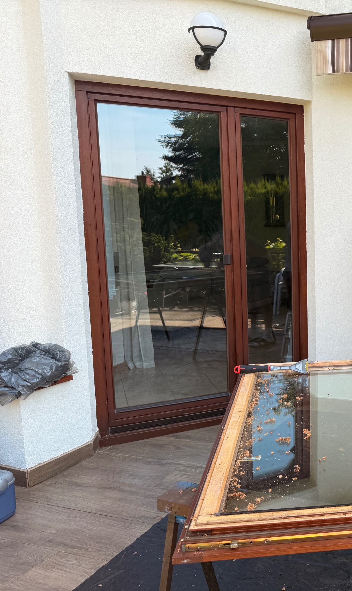 Brązowe dwuskrzydłowe drzwi balkonowe w trakcie renowacji. Widoczna rama okienna na stojaku, narzędzie i worek na odpady. Tło: biała ściana i zieleń.