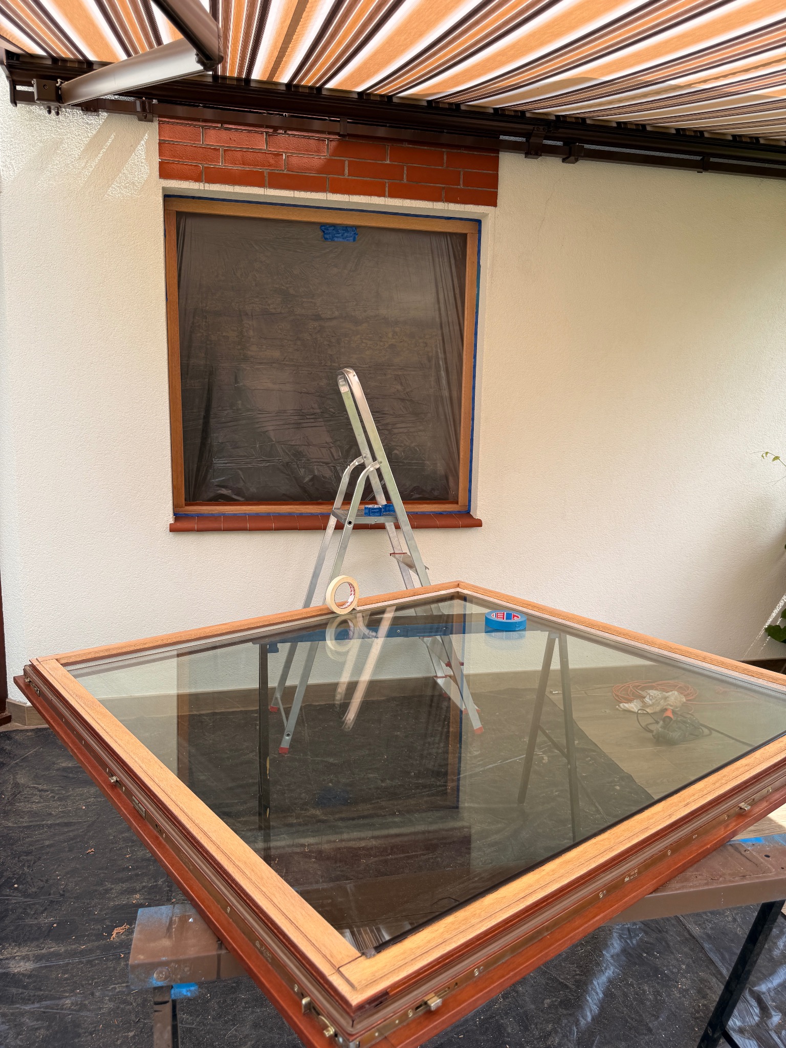 Drewniane okno z szybą przygotowane do montażu, oparte o kozła. W tle okno zabezpieczone folią i drabina. Prace przygotowawcze do instalacji okna.