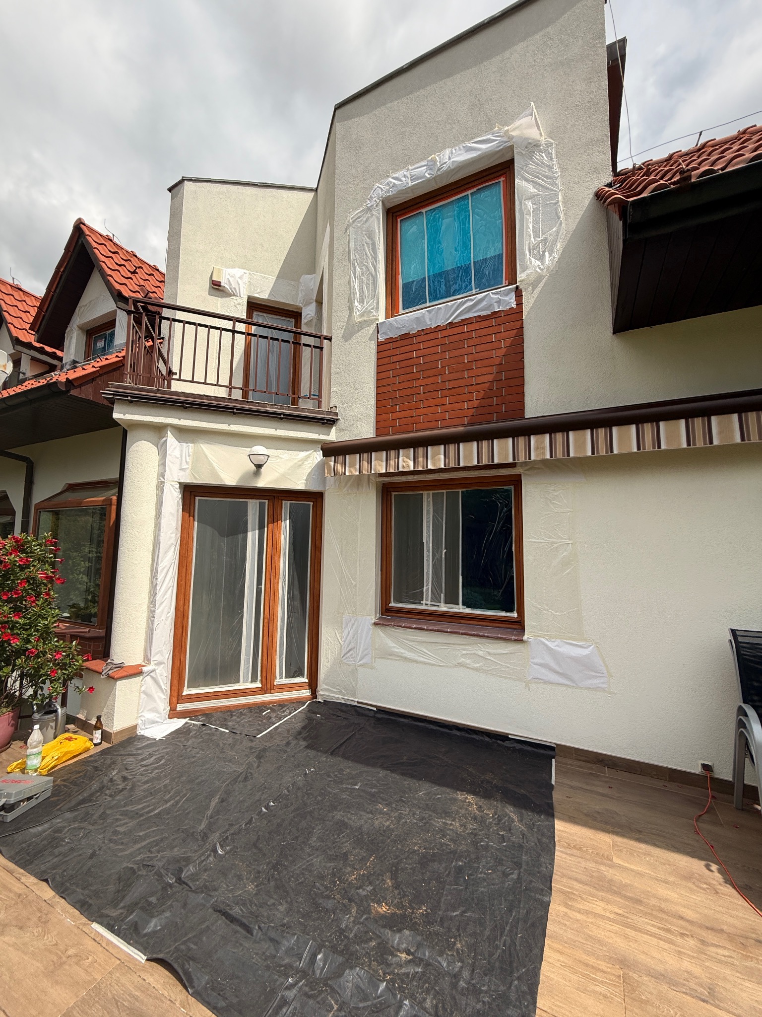 Przygotowania do malowania elewacji: okna i drzwi zabezpieczone folią, podłoże przykryte plandeką. Widoczny fragment domu z czerwonym dachem i balkonem.