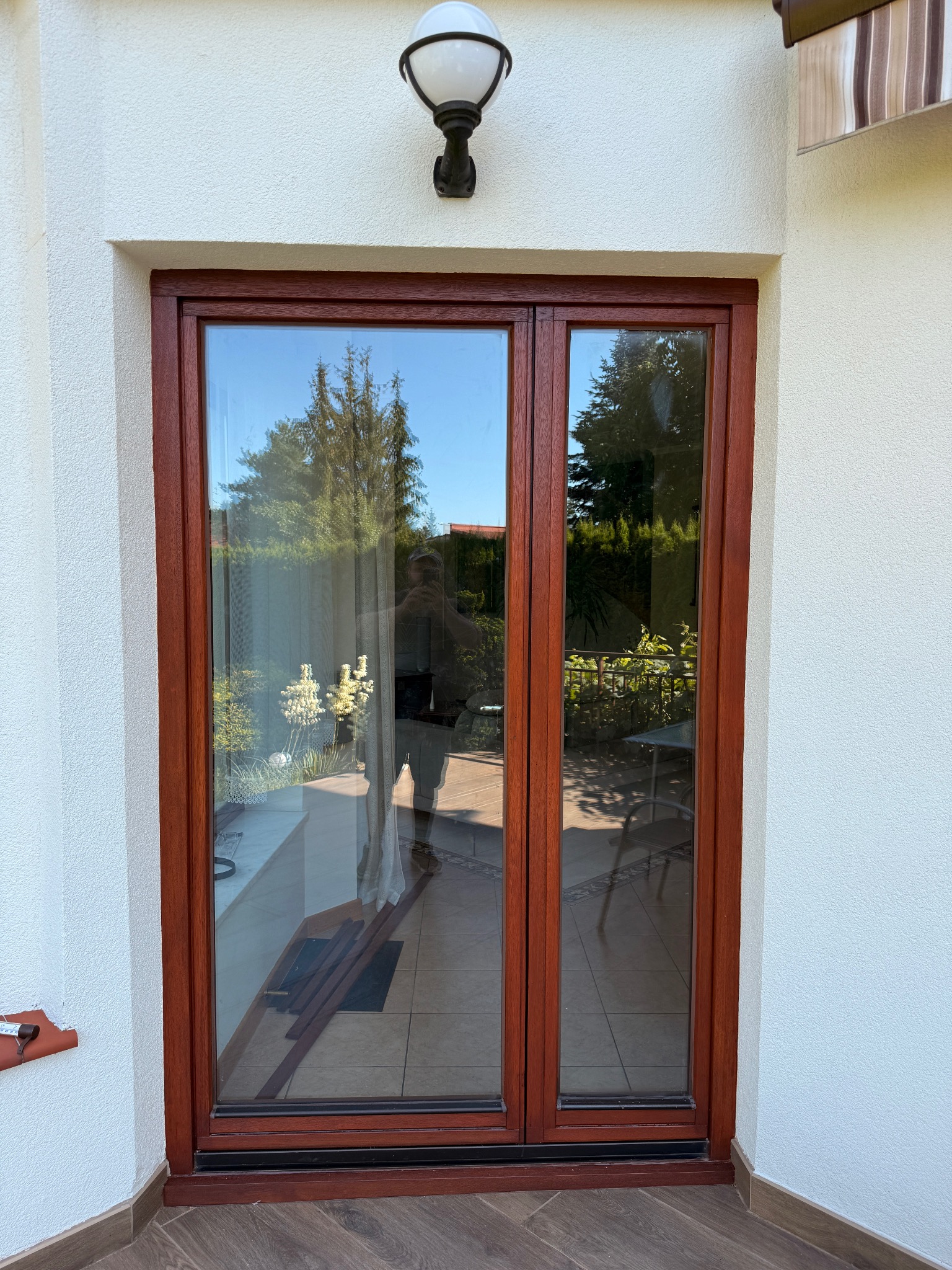 Brązowe, dwuskrzydłowe okno balkonowe z widocznym odbiciem ogrodu i osoby, osadzone w białej elewacji z lampą. Widoczna podłoga z płytek imitujących drewno.