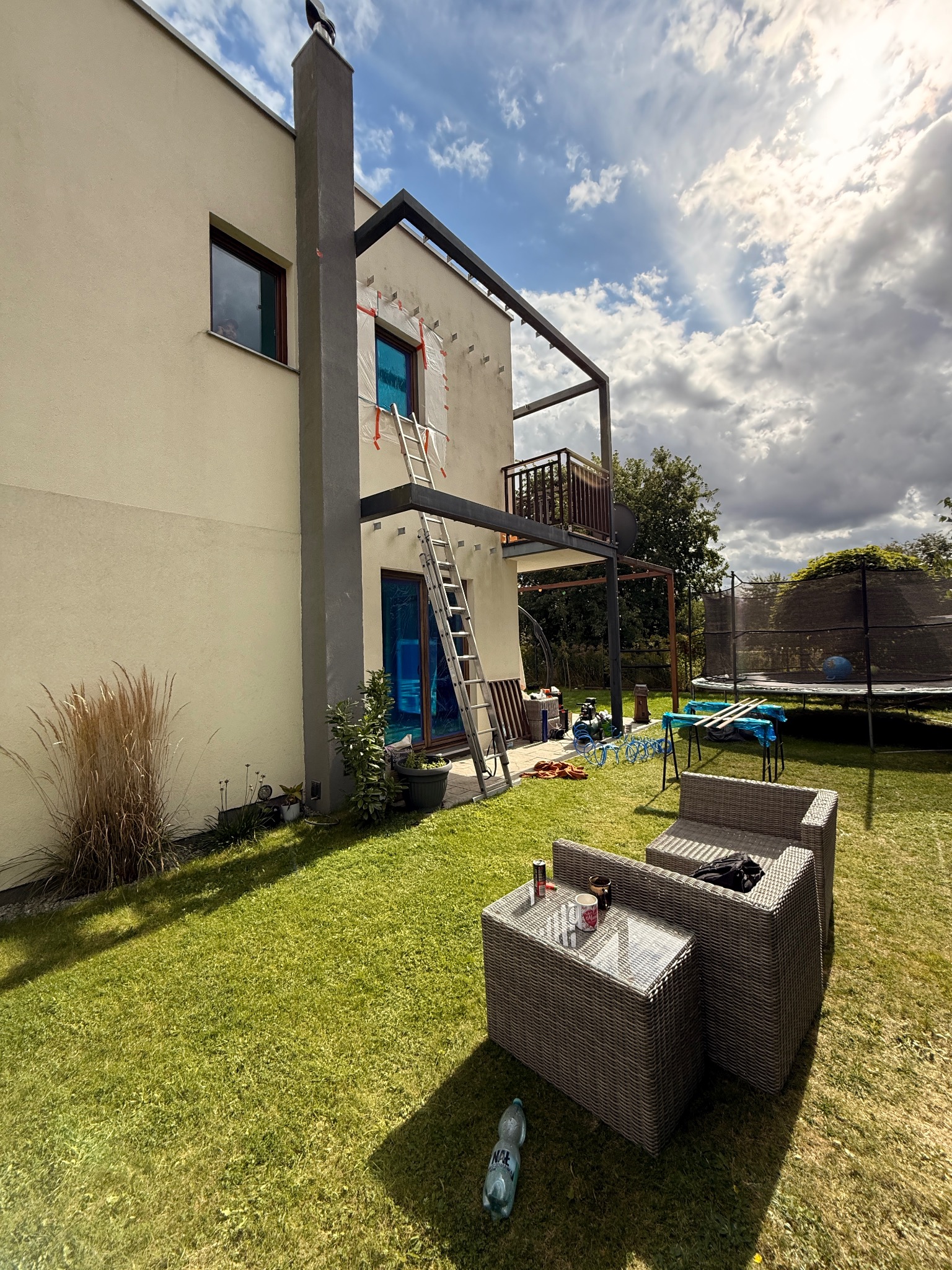 Dwukondygnacyjny dom z oknami zabezpieczonymi folią, drabina oparta o ścianę. Na trawniku narzędzia i meble ogrodowe. Przygotowania do prac remontowych.