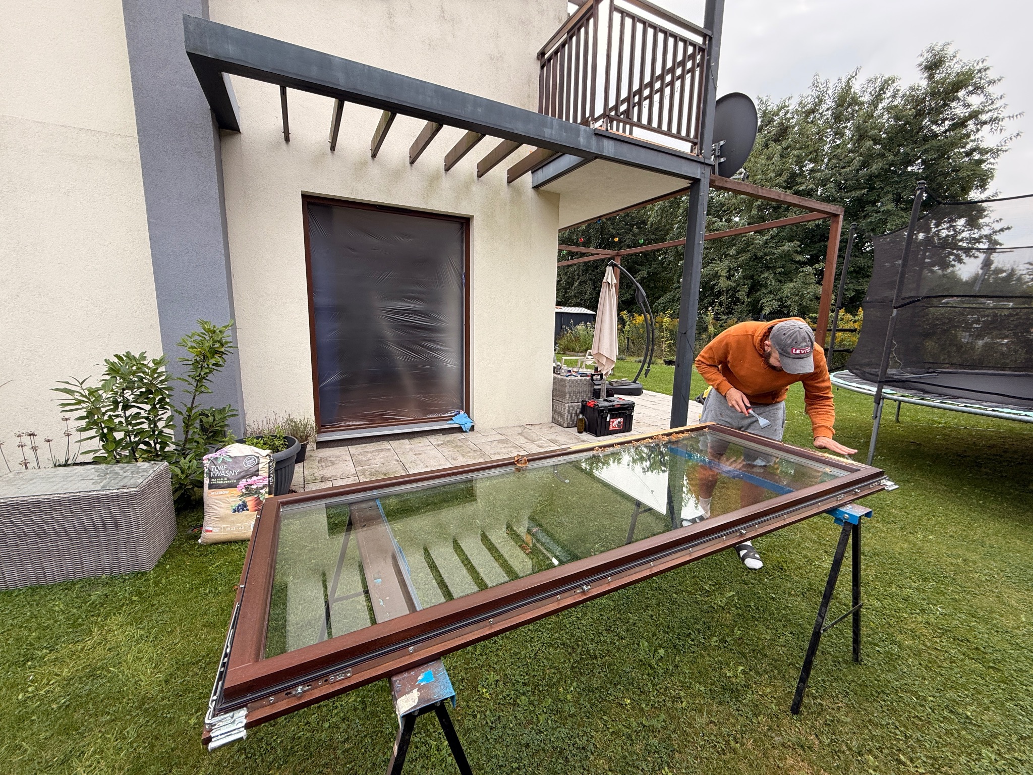 Pracownik w trakcie naprawy okna na zewnątrz budynku. Okno leży na kozłach, widoczne narzędzia i elementy demontażu. W tle trawnik, budynek z balkonem i drzewa.