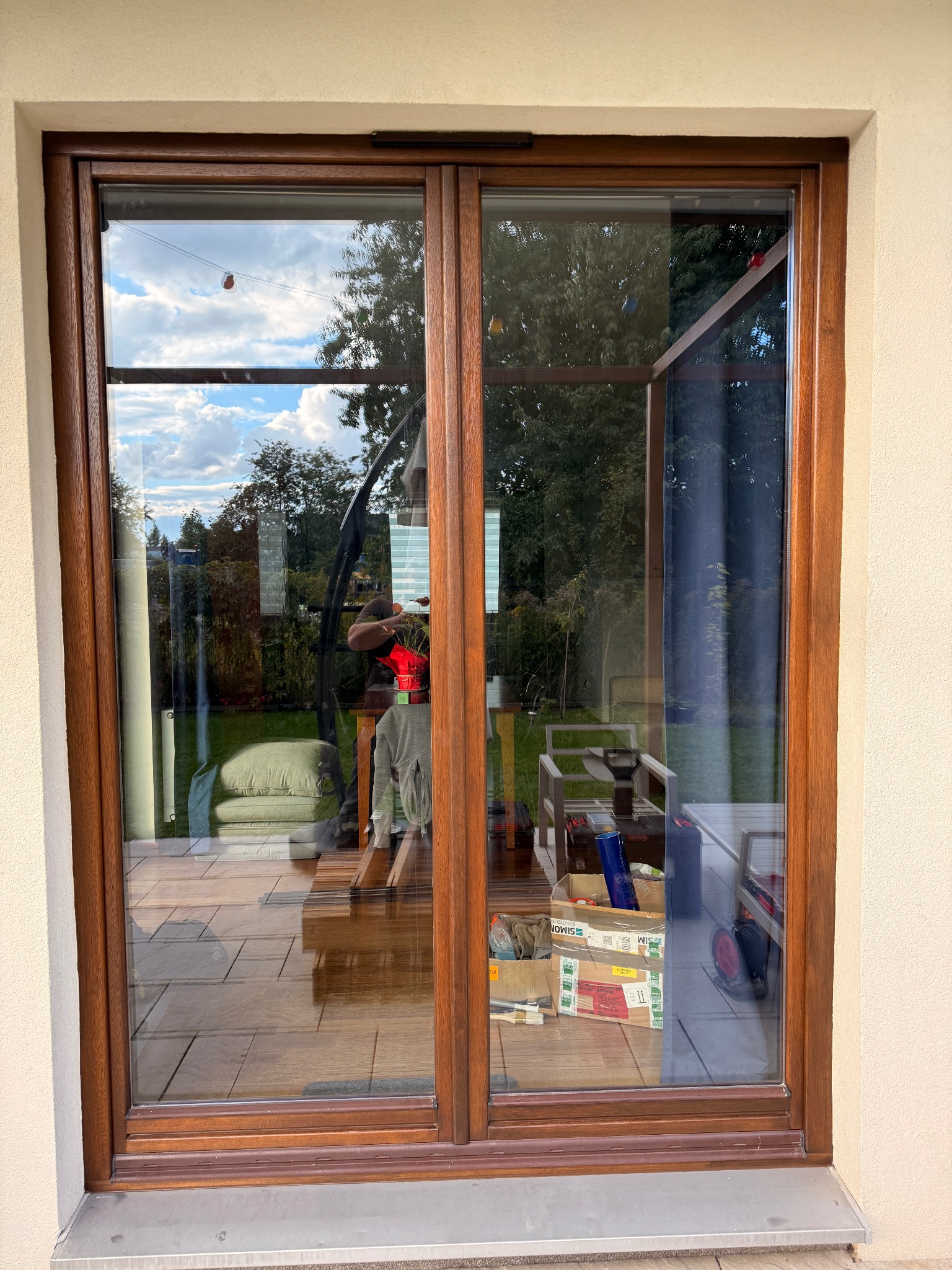 Drewniane, przesuwne okno tarasowe z widokiem na ogród i narzędzia wewnątrz. Okno z ciemnego drewna w jasnej elewacji.