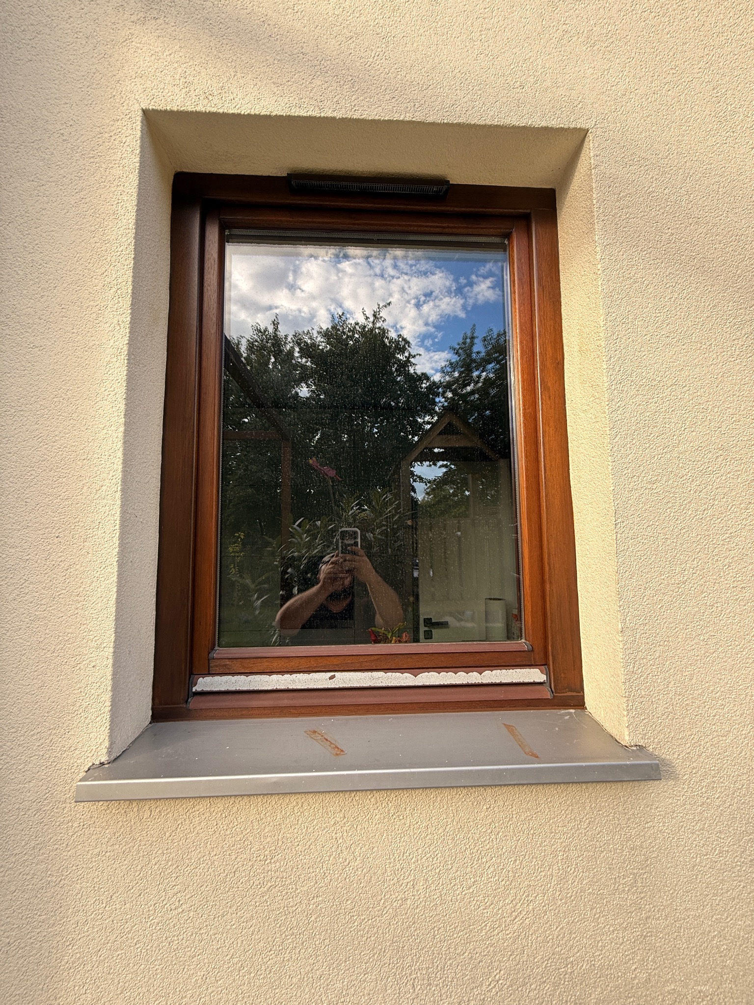 Drewniane okno z widocznym odbiciem osoby robiącej zdjęcie na tle błękitnego nieba i zieleni. Detal elewacji z jasnym, chropowatym tynkiem.