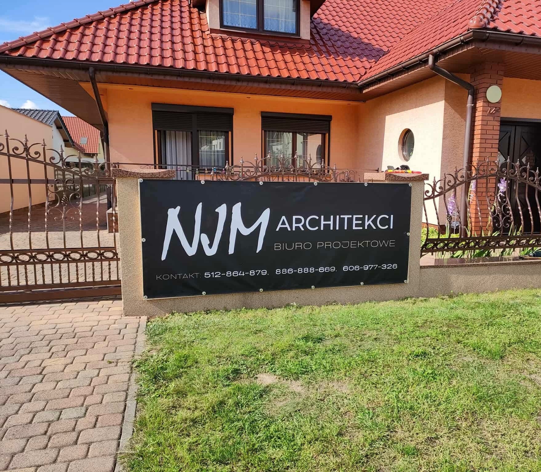 Baner firmy NJM Architekci z numerami telefonów na tle domu jednorodzinnego z czerwoną dachówką i ozdobnym ogrodzeniem.