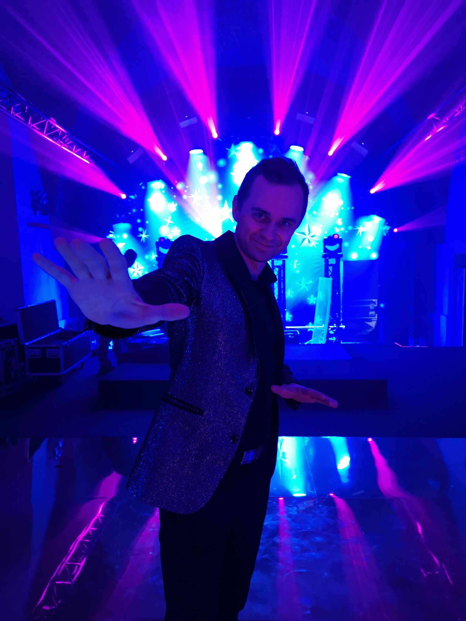 Uśmiechnięty iluzjonista w błyszczącym żakiecie na scenie w niebiesko-różowym oświetleniu, z rozłożonymi rękami, gotowy do występu.