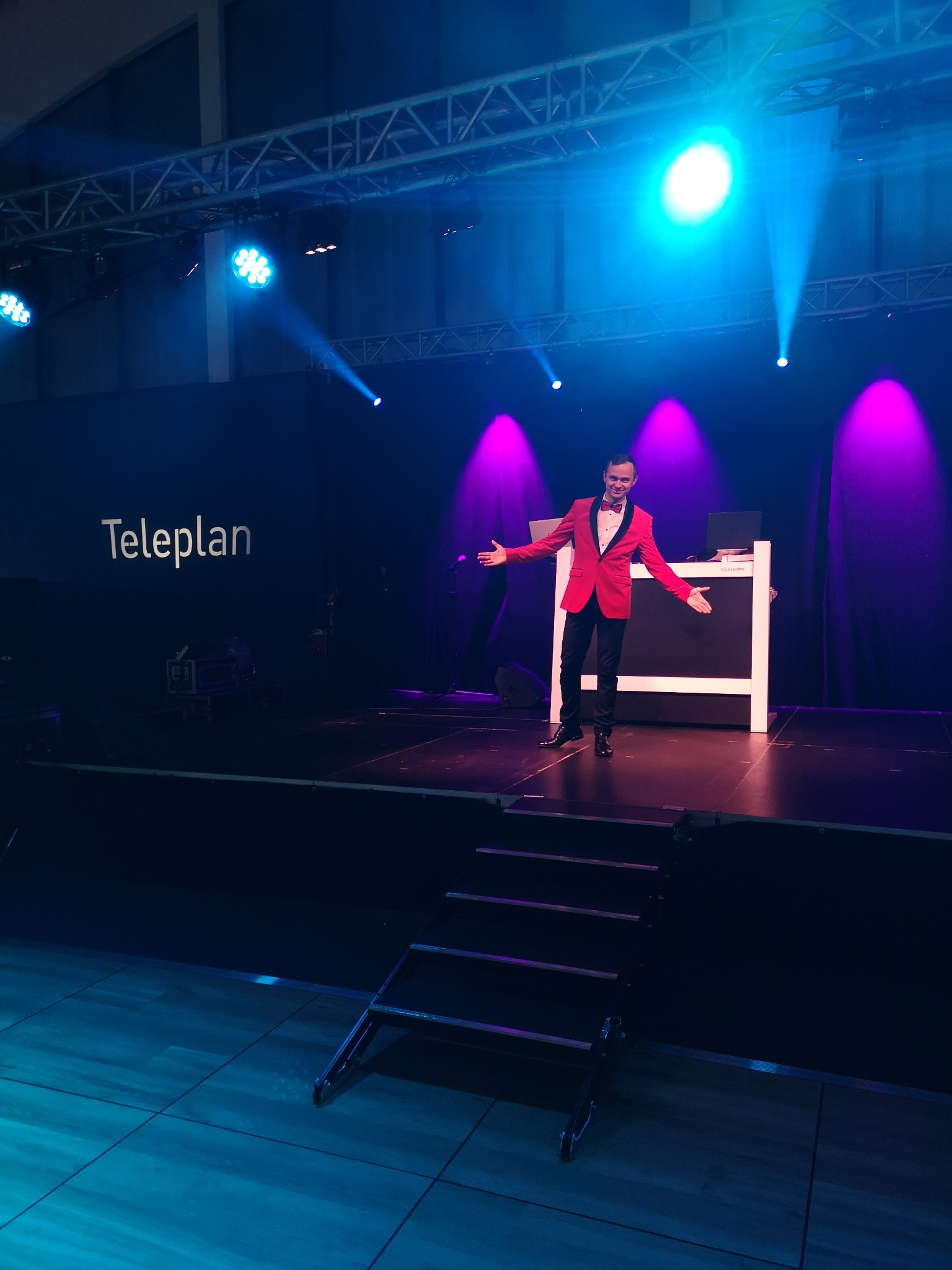 Iluzjonista w czerwonym smokingu na scenie oświetlonej niebieskimi i fioletowymi światłami, z rozłożonymi rękami prezentuje się przed publicznością.