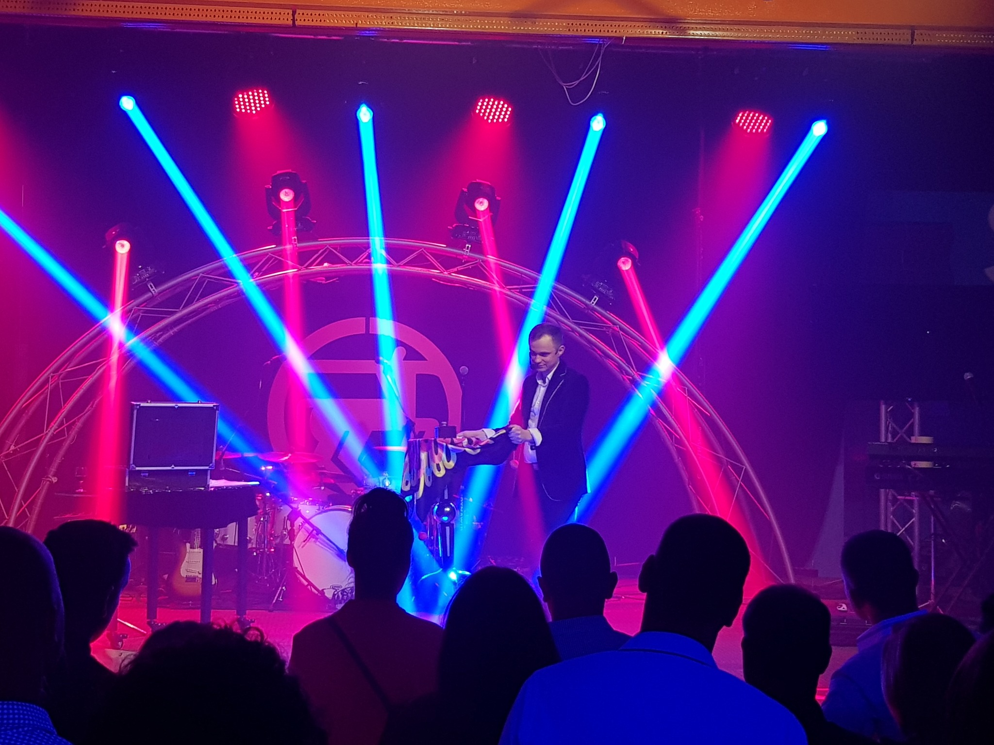 Iluzjonista w trakcie występu na scenie, oświetlony niebieskimi i czerwonymi reflektorami, widoczna publiczność i perkusja w tle.