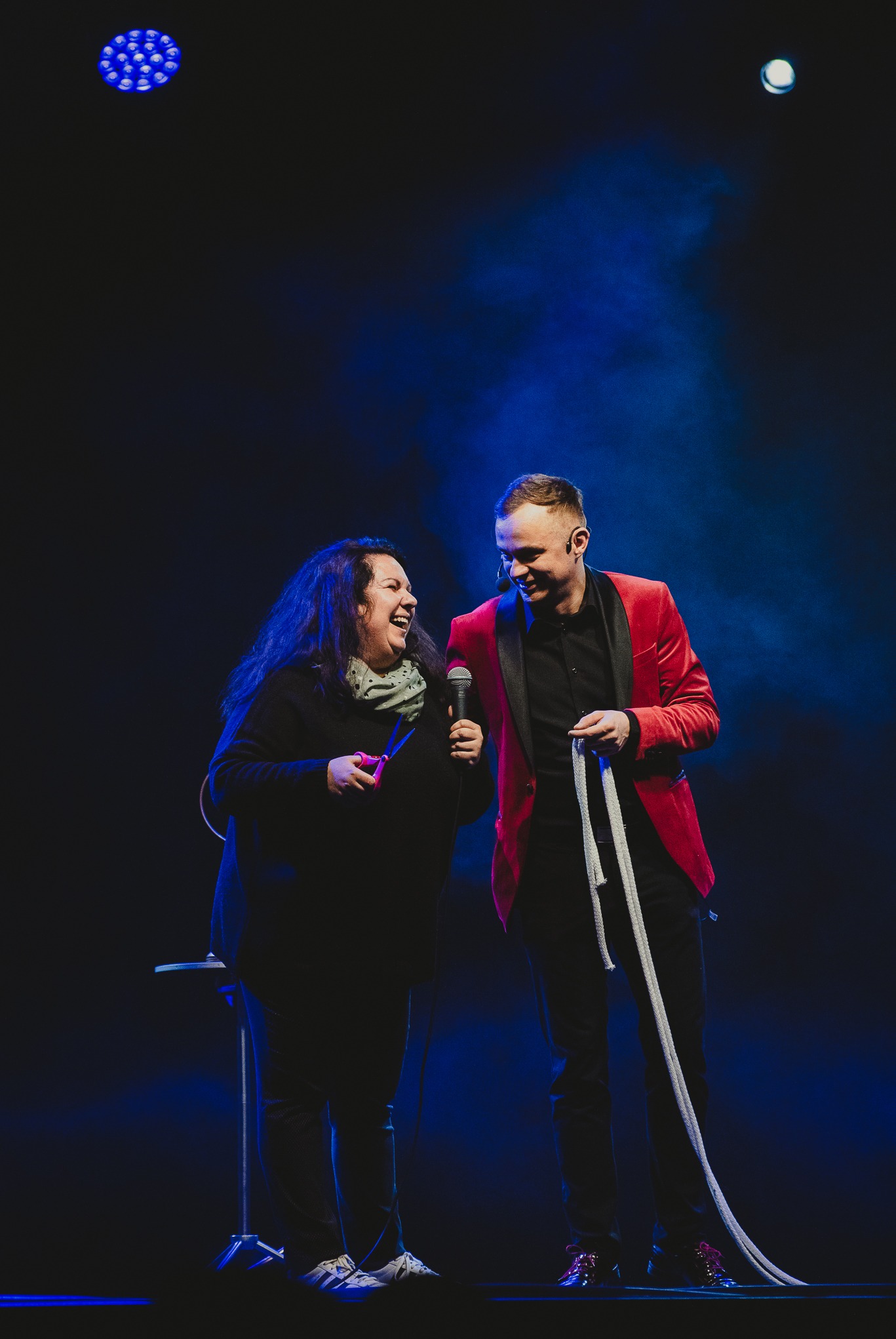 Iluzjonista w czerwonej marynarce prezentuje trik z liną na scenie z kobietą, która trzyma nożyczki i mikrofon, oświetlenie sceniczne.