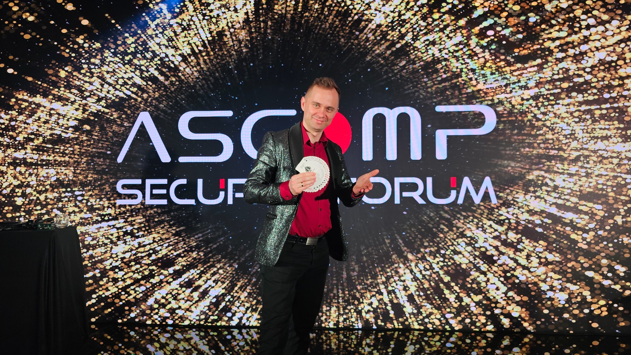 Uśmiechnięty konferansjer w błyszczącej marynarce prezentuje trik z kartami na tle sceny z napisem 'ASCOMP SECURITY FORUM' i efektowną grafiką świetlną.
