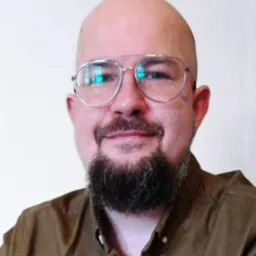 Portret mężczyzny z brodą w okularach, ubranego w brązową koszulę na tle jasnej ściany. W okularach widoczne odbicie okna.