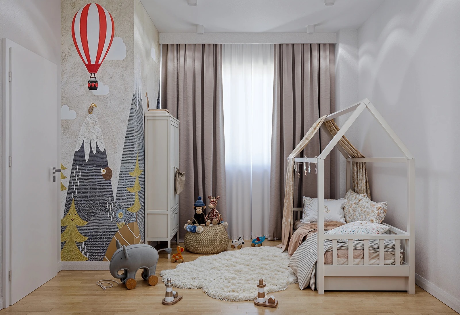 Pokój dziecięcy w neutralnych barwach: łóżko domek, puszysty dywan, ściana z motywem gór i balonu. Jasne drewno, tekstylia w beżach i bielach tworzą przytulną przestrzeń.