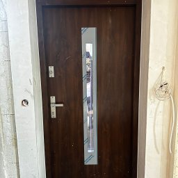 MONT-BUD Dorota Śleziak - Ciemne drzwi wejściowe z szybą, zabezpieczone folią, w surowym otworze. Widoczne kable elektryczne i niedokończone wykończenie ścian.