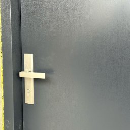 MONT-BUD Dorota Śleziak - Nowoczesne, grafitowe drzwi wejściowe z minimalistyczną, srebrną klamką i zamkiem. Widoczne detale faktury drzwi i żółte zabezpieczenie ościeżnicy.