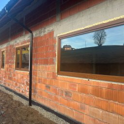 MONT-BUD Dorota Śleziak - Montaż okien w surowym budynku z cegły. Widoczne brązowe ramy okienne i pianka montażowa. W szybach odbija się krajobraz.