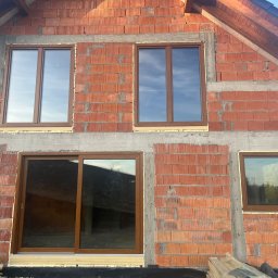 MONT-BUD Dorota Śleziak - Surowy budynek z brązowymi oknami PCV w trakcie montażu, widoczne pustaki i betonowe wzmocnienia, odbicie nieba w szybach.