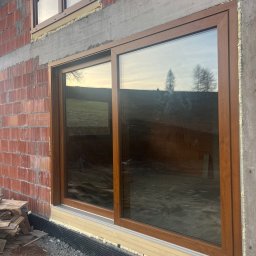 MONT-BUD Dorota Śleziak - Brązowe okno przesuwne, zamontowane w surowym murze z cegły, z widoczną izolacją i odblaskiem krajobrazu w szybie. Widoczne detale montażu.