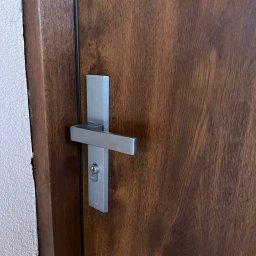 MONT-BUD Dorota Śleziak - Ciemnobrązowe drzwi z drewnianą fakturą i nowoczesną, srebrną klamką z zamkiem, widoczne na tle jasnej ściany.