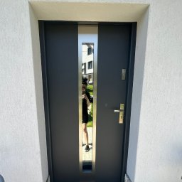 MONT-BUD Dorota Śleziak - Grafitowe drzwi wejściowe z metalową wstawką, widoczne na tle białej, chropowatej elewacji i kamiennej podmurówki. W lustrzanym panelu odbija się osoba i otoczenie.