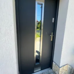 Nowoczesne, grafitowe drzwi wejściowe z przeszkleniem, stalowe wykończenia, na tle białej ściany z fakturą i szarej kostki brukowej. Widok z bliska, lekki kąt.