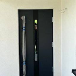 MONT-BUD Dorota Śleziak - Nowoczesne, czarne drzwi zewnętrzne z wąskim przeszkleniem pośrodku, częściowo zabezpieczone folią bąbelkową. Widoczne białe ściany i fragment osoby w odbiciu.