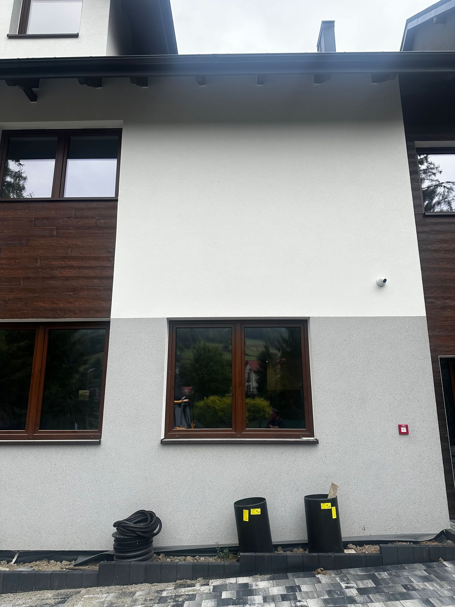 Elewacja domu z brązowymi oknami i drewnianą okładziną. Widoczne dwa czarne słupki i zwinięty wąż. Ujęcie z dołu, pochmurne niebo.