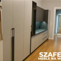 Jasnoszara zabudowa kuchenna z czarnymi uchwytami, widok z perspektywy wejścia, drewniana podłoga, lustro w tle, minimalistyczny design.