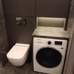 Szara zabudowa meblowa w łazience z podświetleniem, obejmująca toaletę i pralkę Samsung. Marmurowe płytki podłogowe. Ujęcie z dołu.