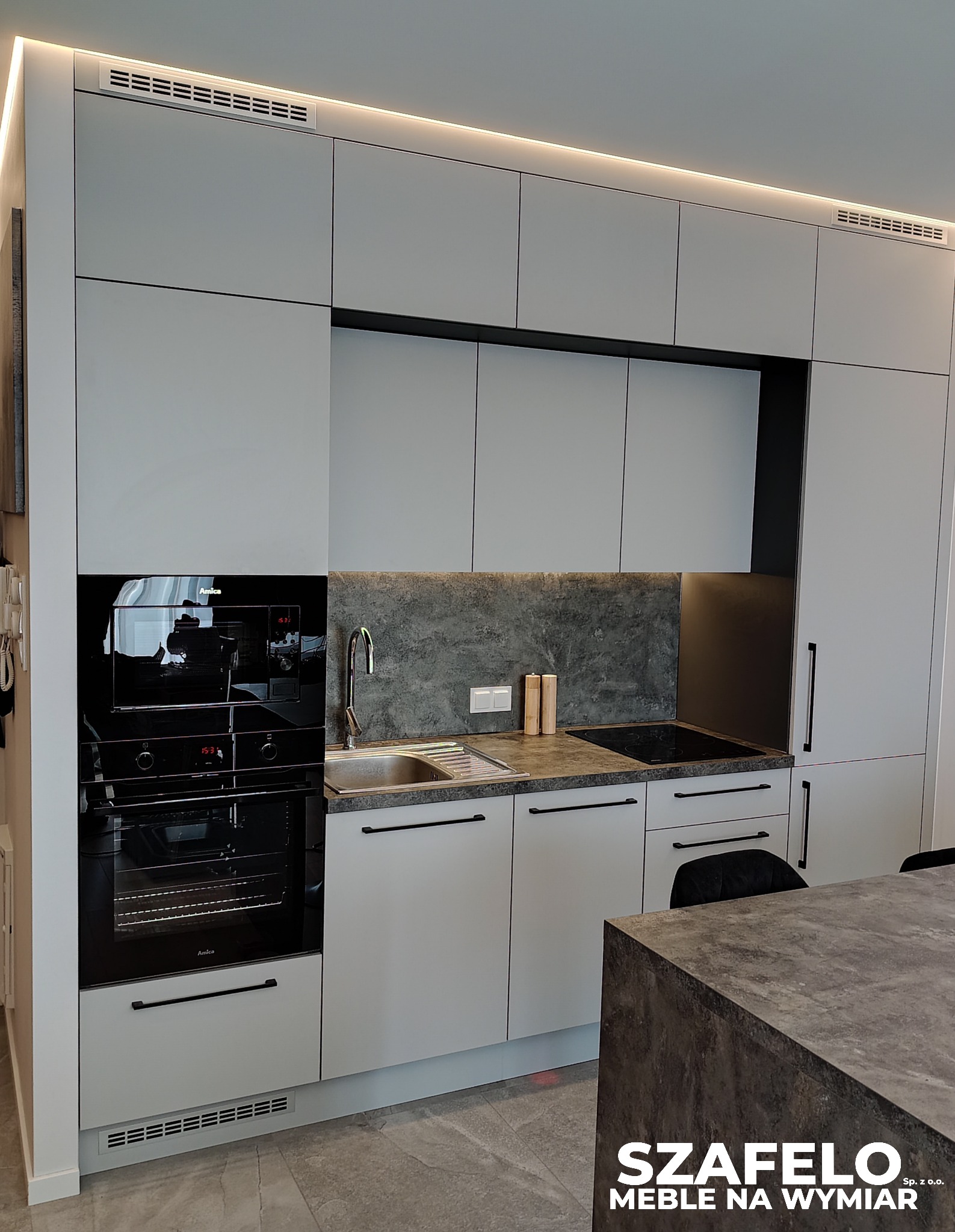 Nowoczesna kuchnia z szarymi frontami, czarnymi detalami i blatem imitującym beton. Oświetlenie LED podkreśla minimalistyczny design i dodaje elegancji wnętrzu.