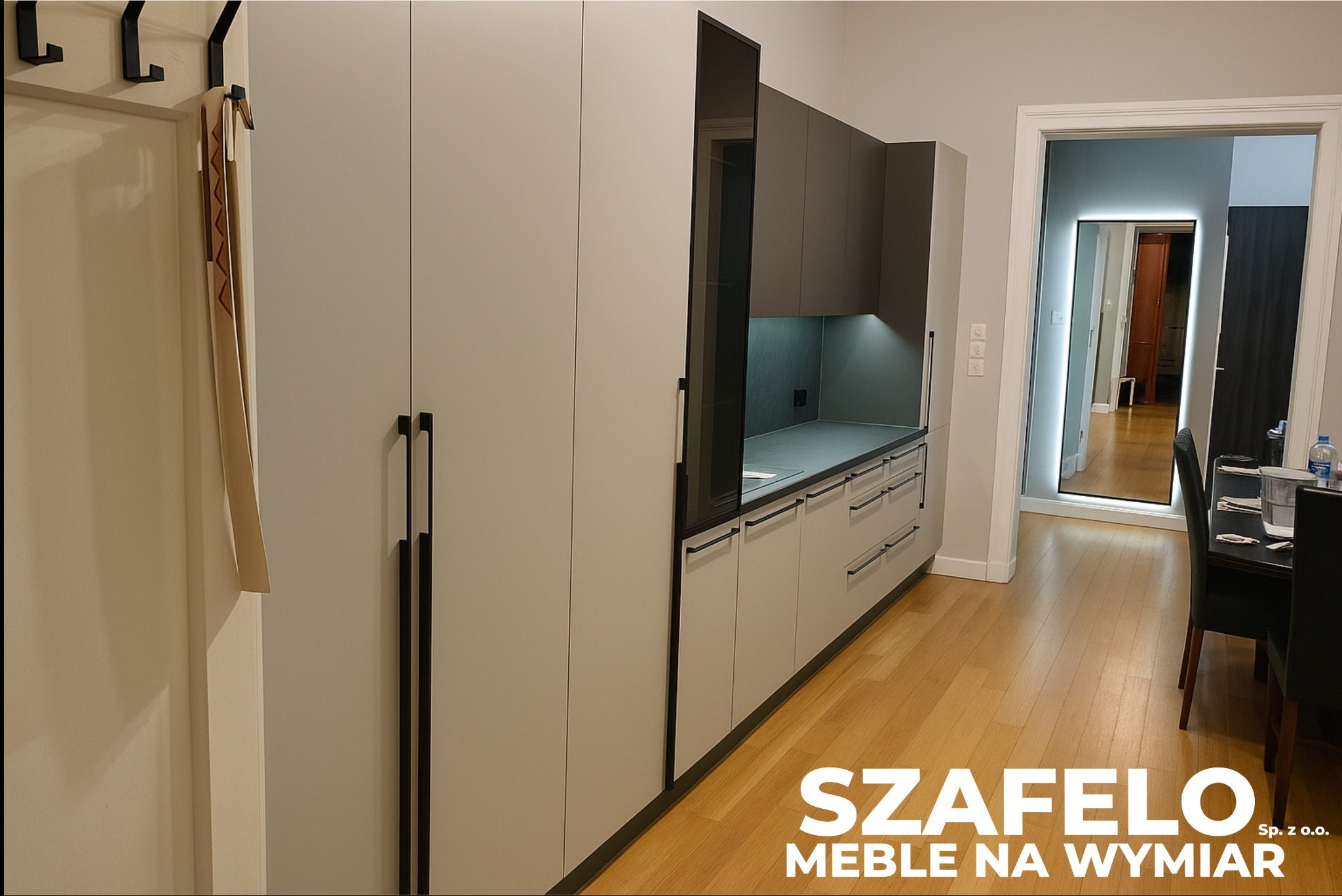 Jasnoszara zabudowa kuchenna z czarnymi uchwytami, widok z perspektywy wejścia, drewniana podłoga, lustro w tle, minimalistyczny design.