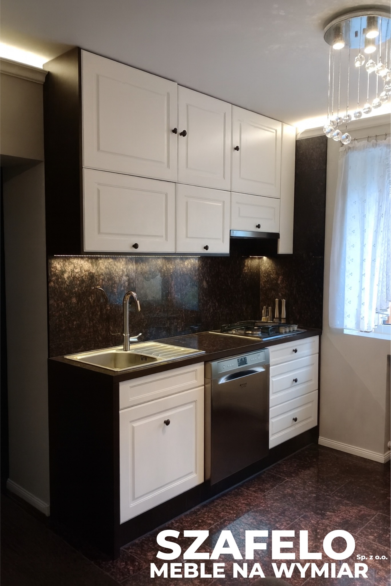 Nowoczesna kuchnia z białymi szafkami i ciemnym blatem, oświetlona listwami LED, z widocznym zlewem, płytą gazową i zmywarką. Kryształowy żyrandol dopełnia aranżację.