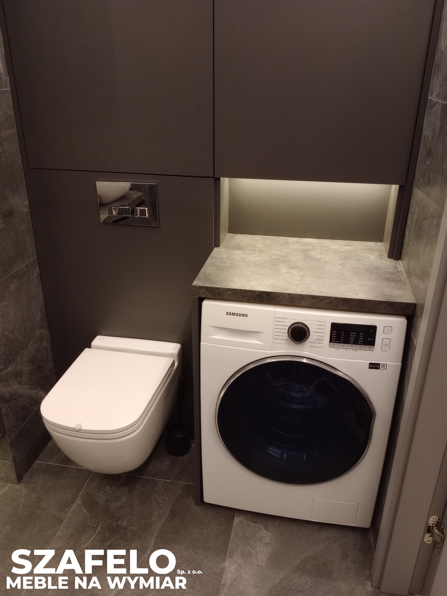 Szara zabudowa meblowa w łazience z podświetleniem, obejmująca toaletę i pralkę Samsung. Marmurowe płytki podłogowe. Ujęcie z dołu.