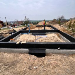 Cameleon firma budowlana Patryk Ratajczak - Etap budowy: fundamenty domu z izolacją, widoczny pracownik w odblaskowej kamizelce, rury instalacyjne. Tło: teren budowy, niebo.