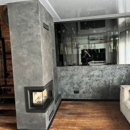 Cameleon firma budowlana Patryk Ratajczak - Nowoczesny salon z kominkiem obłożonym szarym, teksturowanym tynkiem dekoracyjnym, widok na schody z drewnianymi stopniami i przeszkloną zabudowę ścienną.