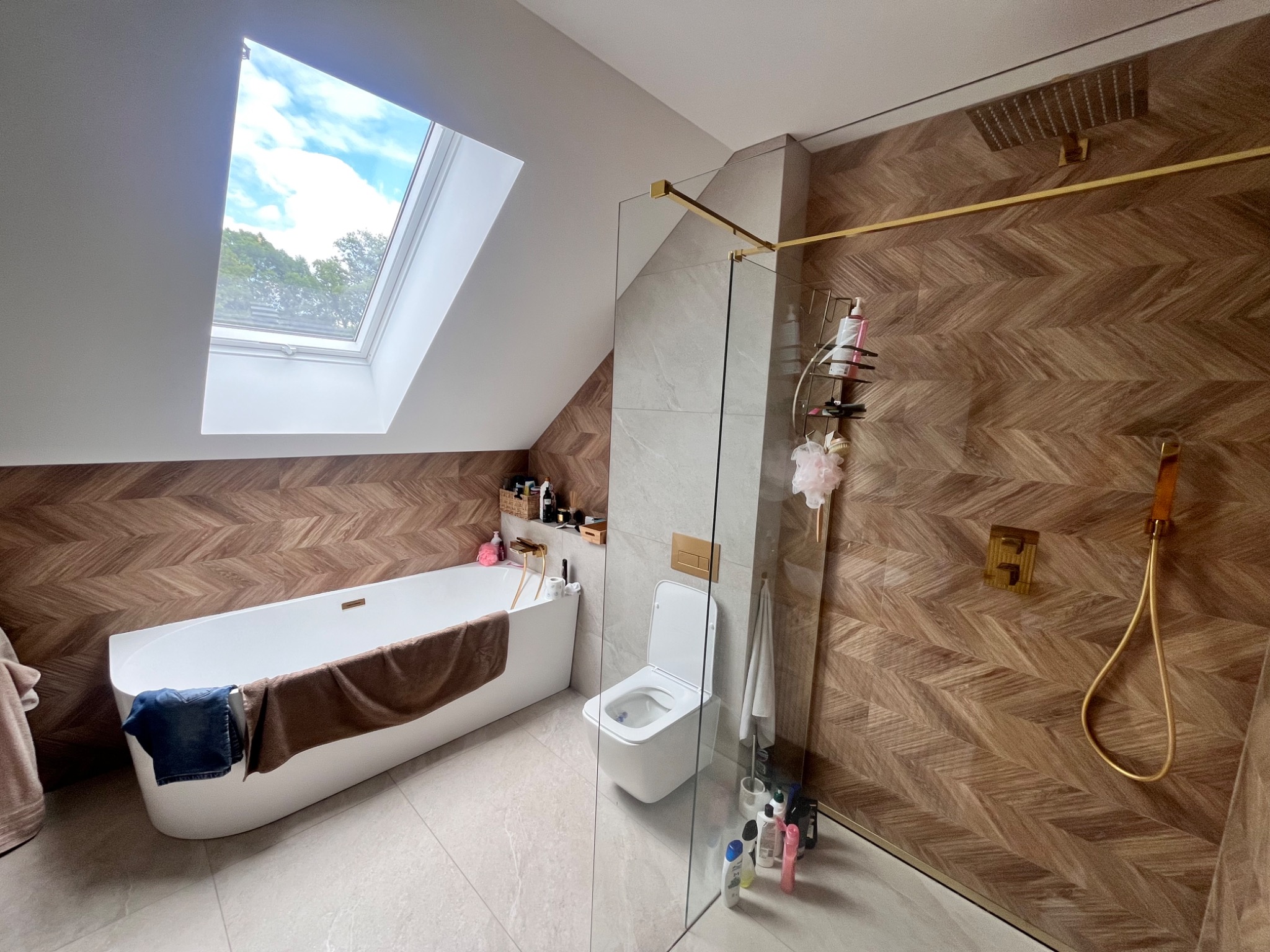 Nowoczesna łazienka na poddaszu z oknem dachowym. Drewnopodobne płytki ułożone w jodełkę, biała wanna i toaleta, kabina prysznicowa ze złotymi elementami.