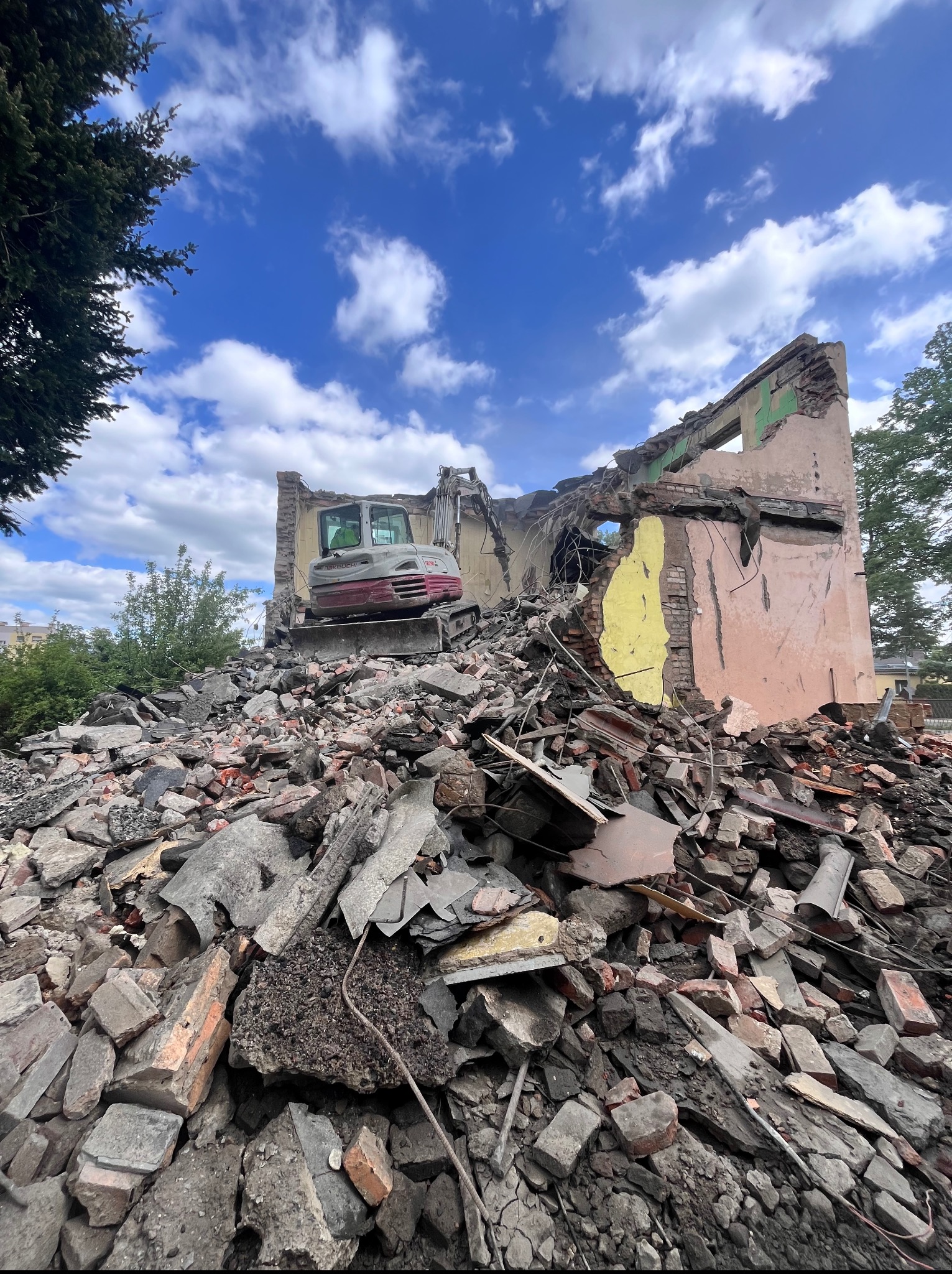 Rozbiórka budynku z użyciem koparki na tle błękitnego nieba z chmurami. Gruz i fragmenty ścian w pierwszym planie. Widok z dołu podkreśla skalę zniszczeń.