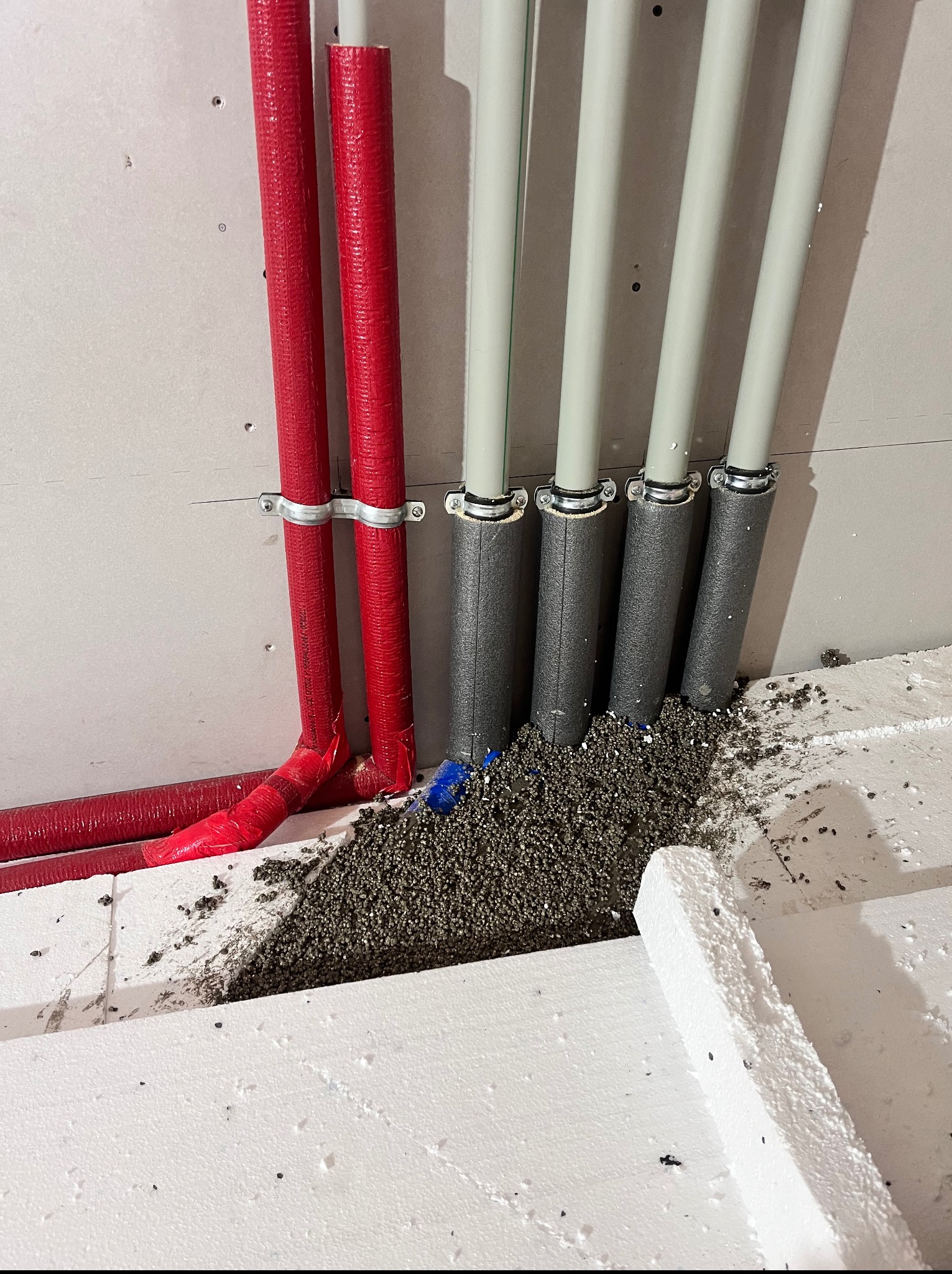 Rury CO i wodne w izolacji, mocowane do ściany z płyt karton-gips, z widocznym styropianem i granulatem izolacyjnym na podłodze.