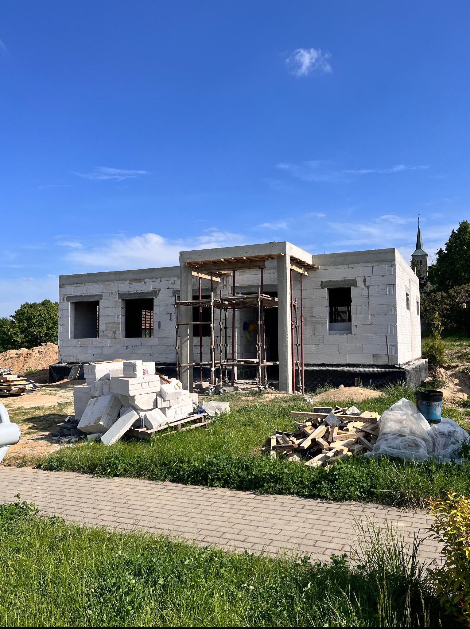 Budowa domu jednorodzinnego w stanie surowym otwartym, widoczne białe bloczki, rusztowanie, materiały budowlane, trawa i drzewa w tle, błękitne niebo.