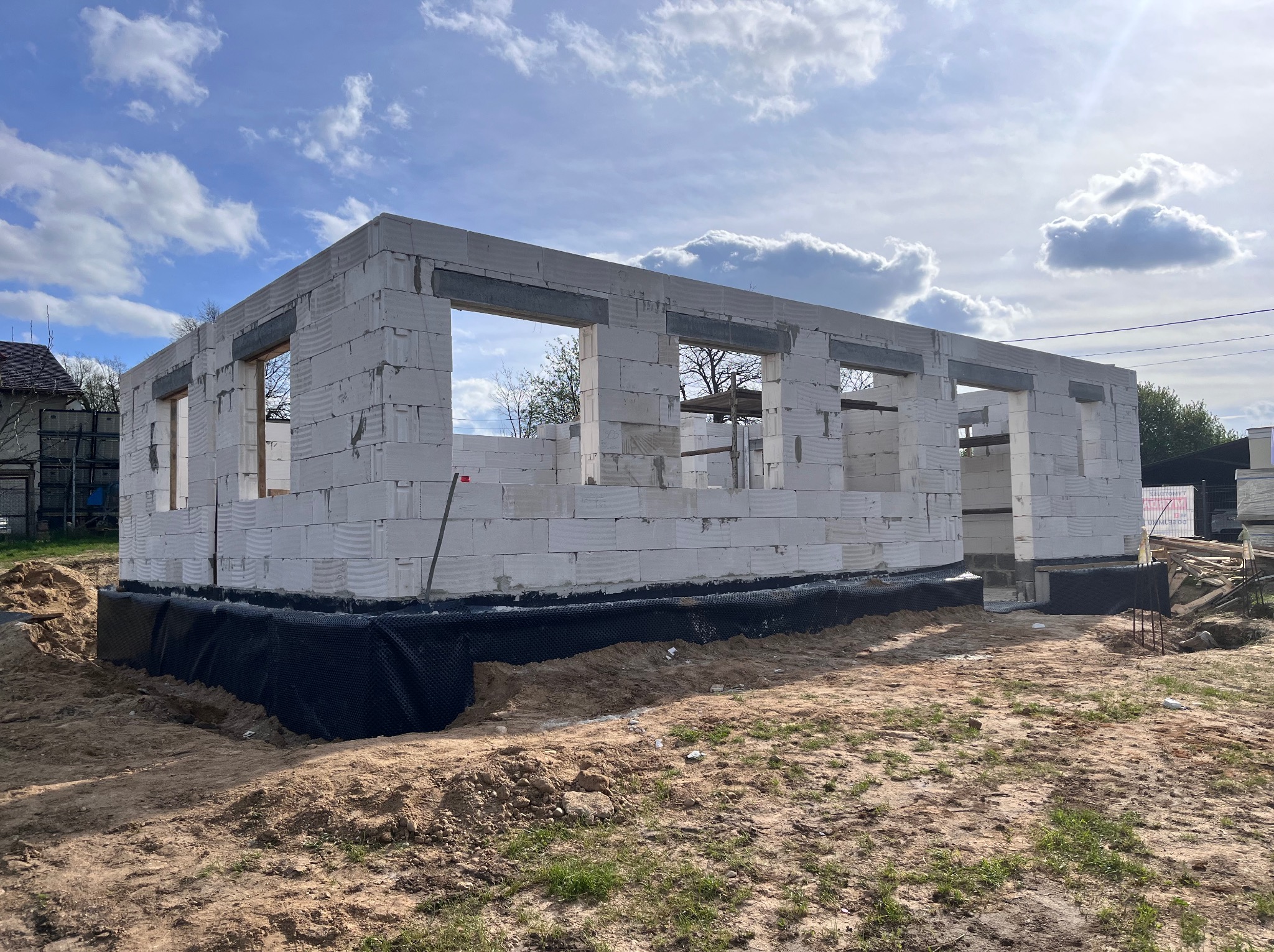 Wznoszenie ścian parterowego domu z białych bloczków, widoczne okna i fundamenty z izolacją, na zewnątrz plac budowy w słoneczny dzień.
