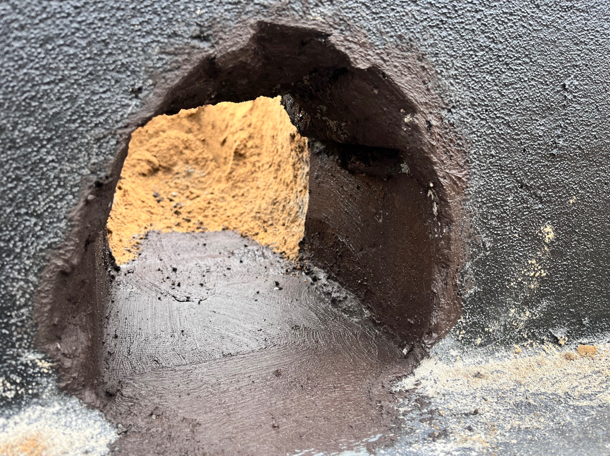 Otwór w ścianie z widoczną warstwą zaprawy i piasku. Świeżo nałożona, ciemna masa uszczelniająca otacza otwór, kontrastując z jasnym podłożem.
