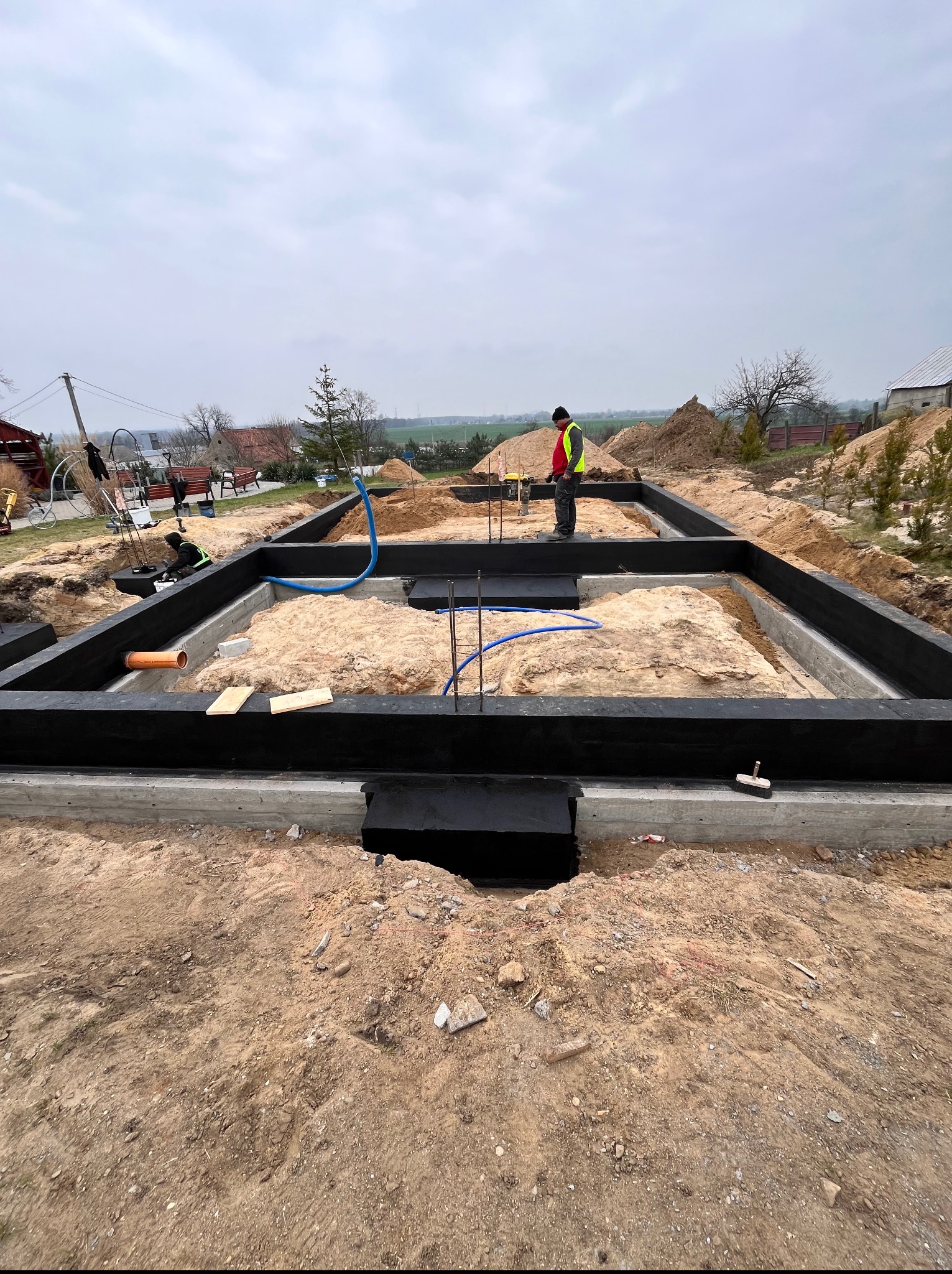 Etap budowy: fundamenty domu z izolacją, widoczny pracownik w odblaskowej kamizelce, rury instalacyjne. Tło: teren budowy, niebo.