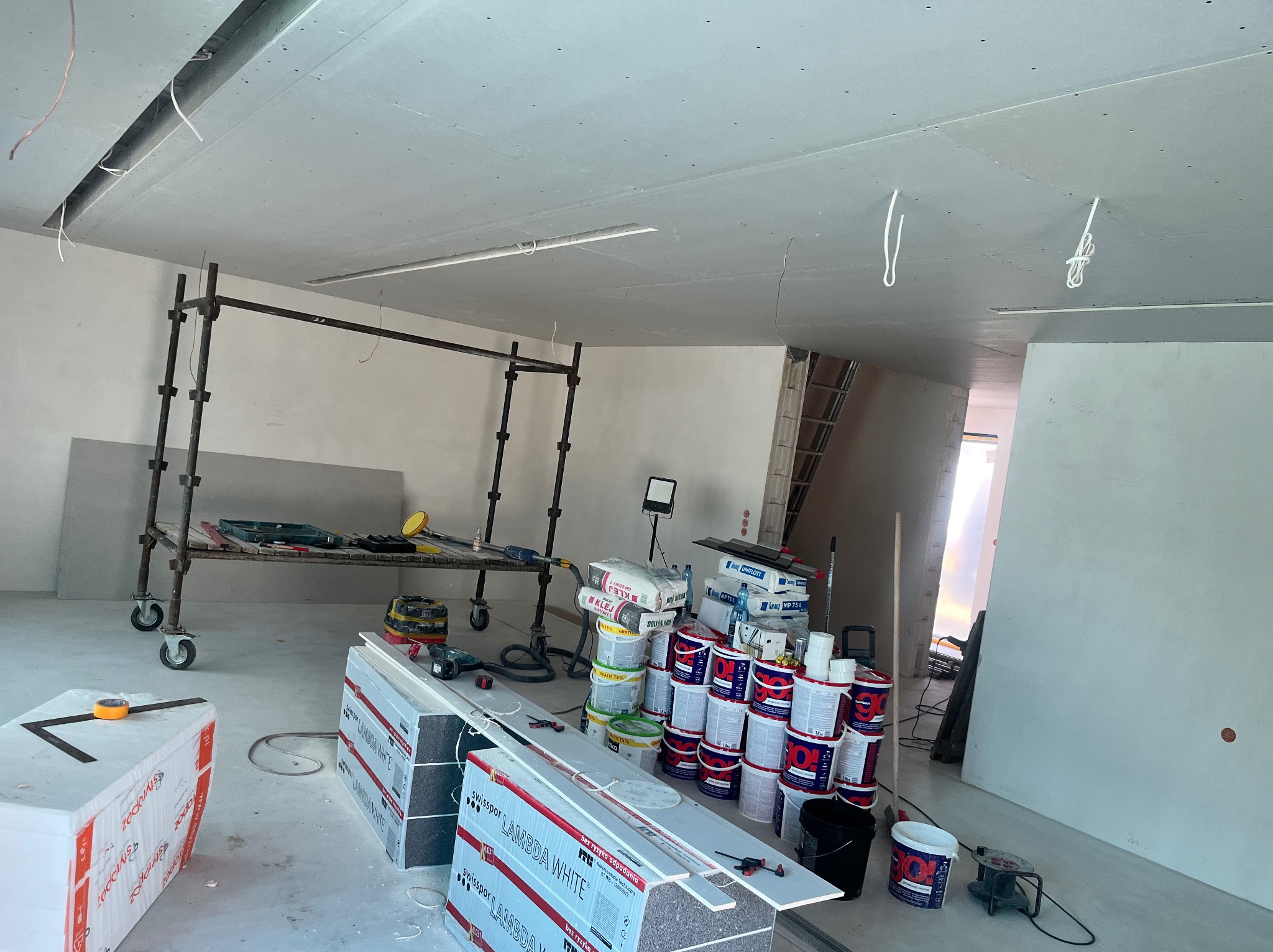 Wnętrze w trakcie prac wykończeniowych: rusztowanie, materiały budowlane (styropian, farby, kleje) i narzędzia. Sufit z płyt kartonowo-gipsowych.