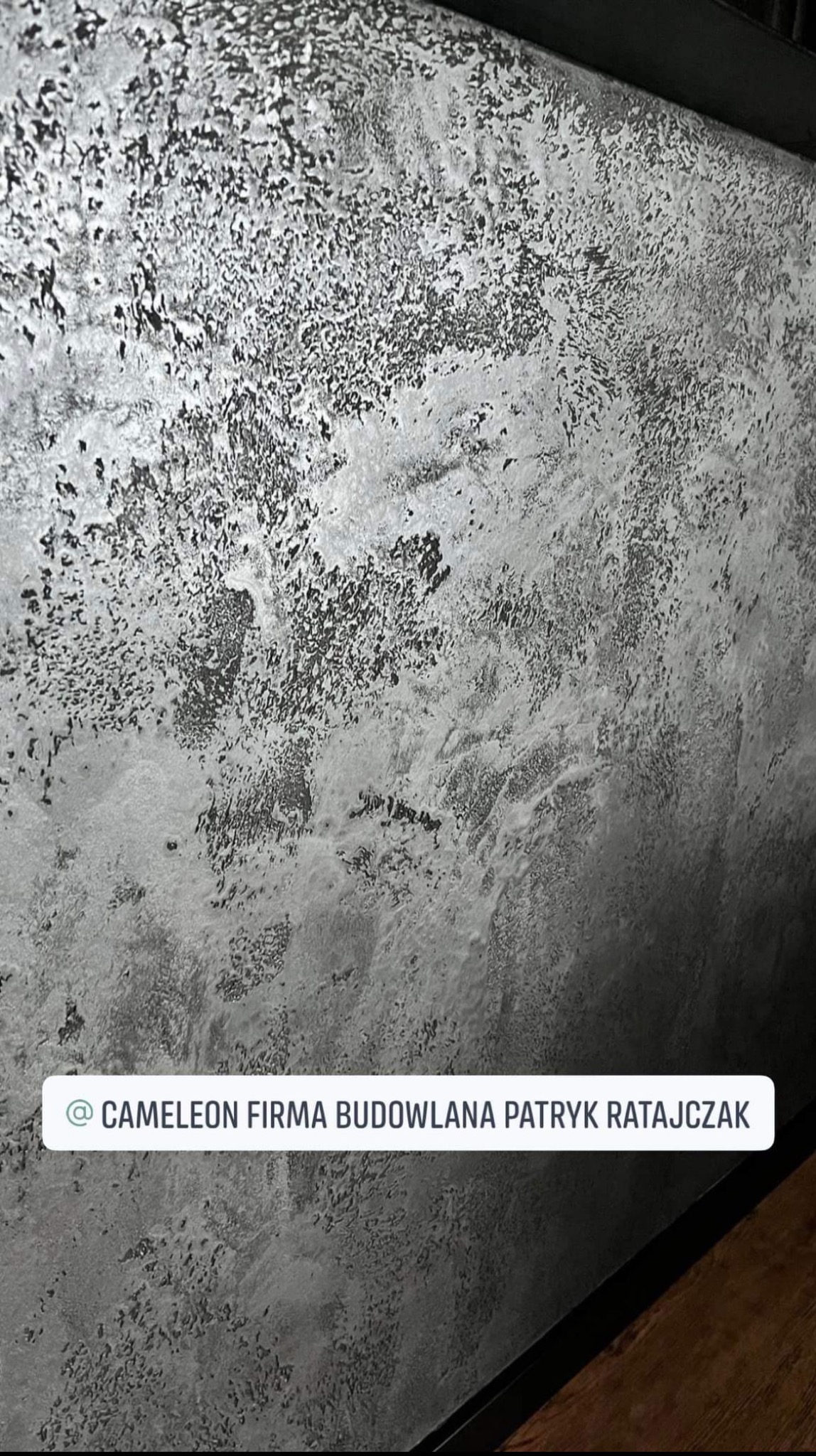 Szara ściana z dekoracyjną fakturą, imitującą kamień lub beton. Widoczny fragment podłogi i sufitu. Nieregularne wzory tworzą unikalny efekt wizualny.