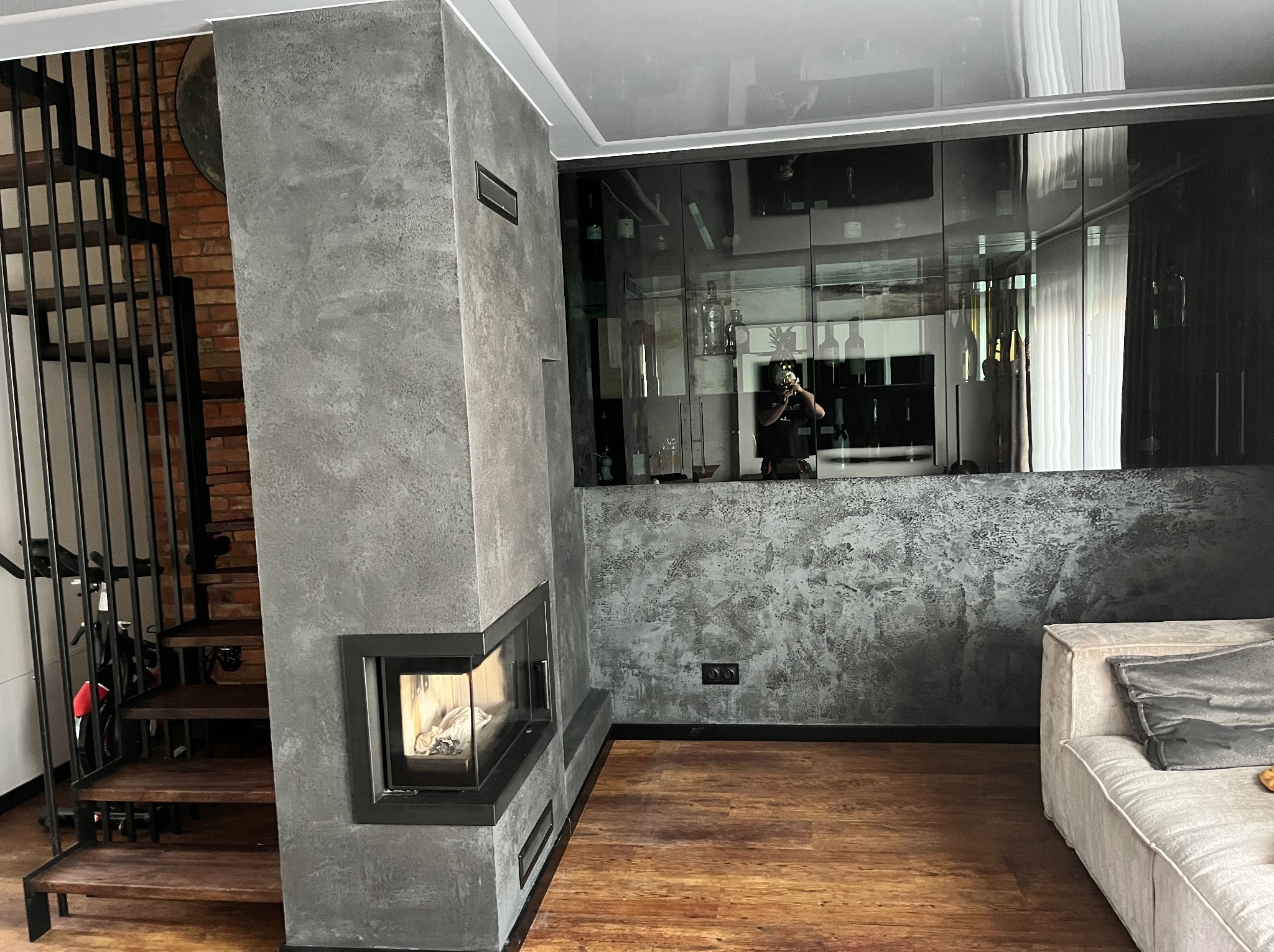 Nowoczesny salon z kominkiem obłożonym szarym, teksturowanym tynkiem dekoracyjnym, widok na schody z drewnianymi stopniami i przeszkloną zabudowę ścienną.