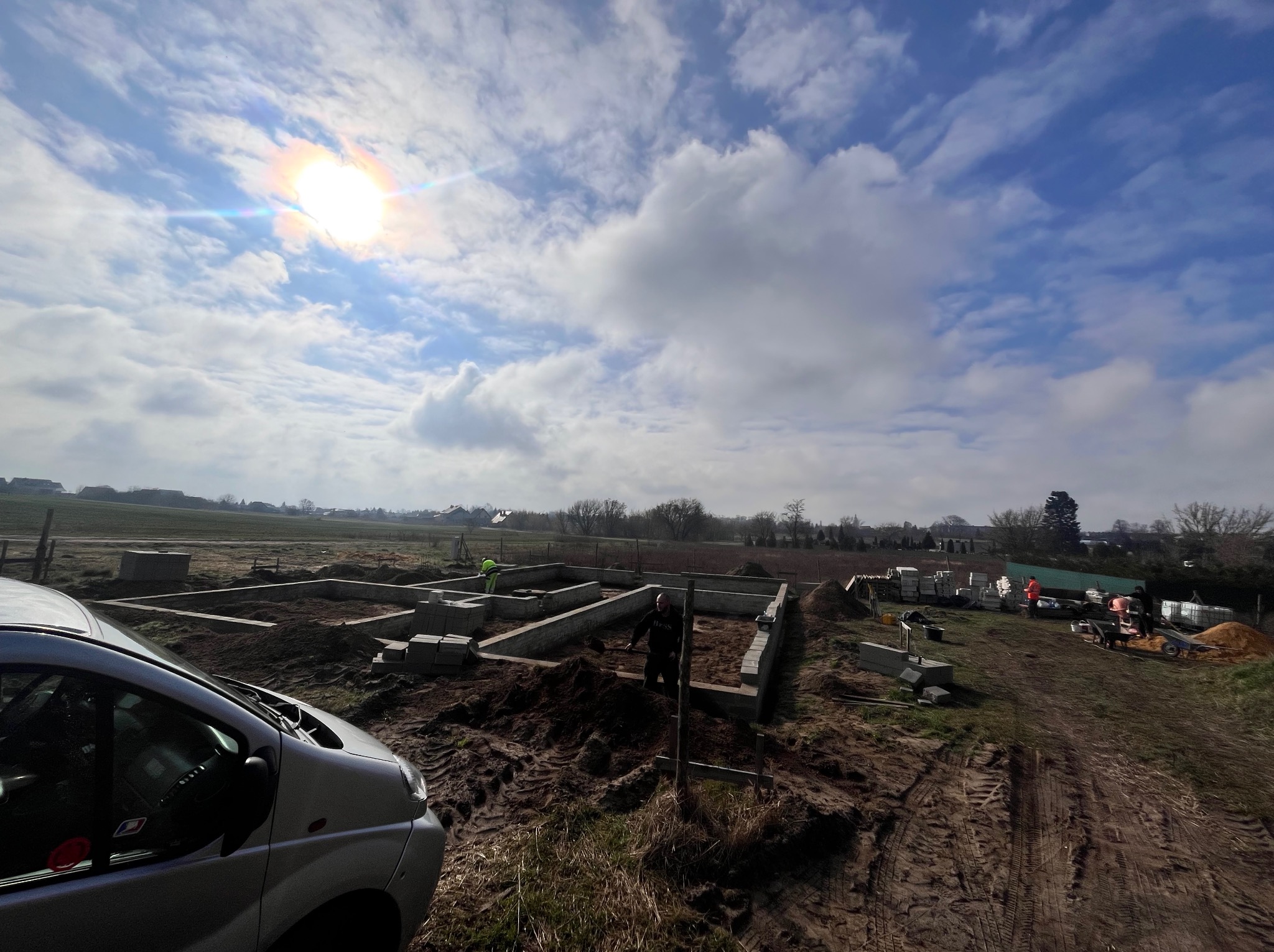 Budowa fundamentów domu jednorodzinnego z betonu komórkowego, widok z perspektywy ziemi, częściowo zasłonięty samochodem dostawczym, pracownicy w tle.