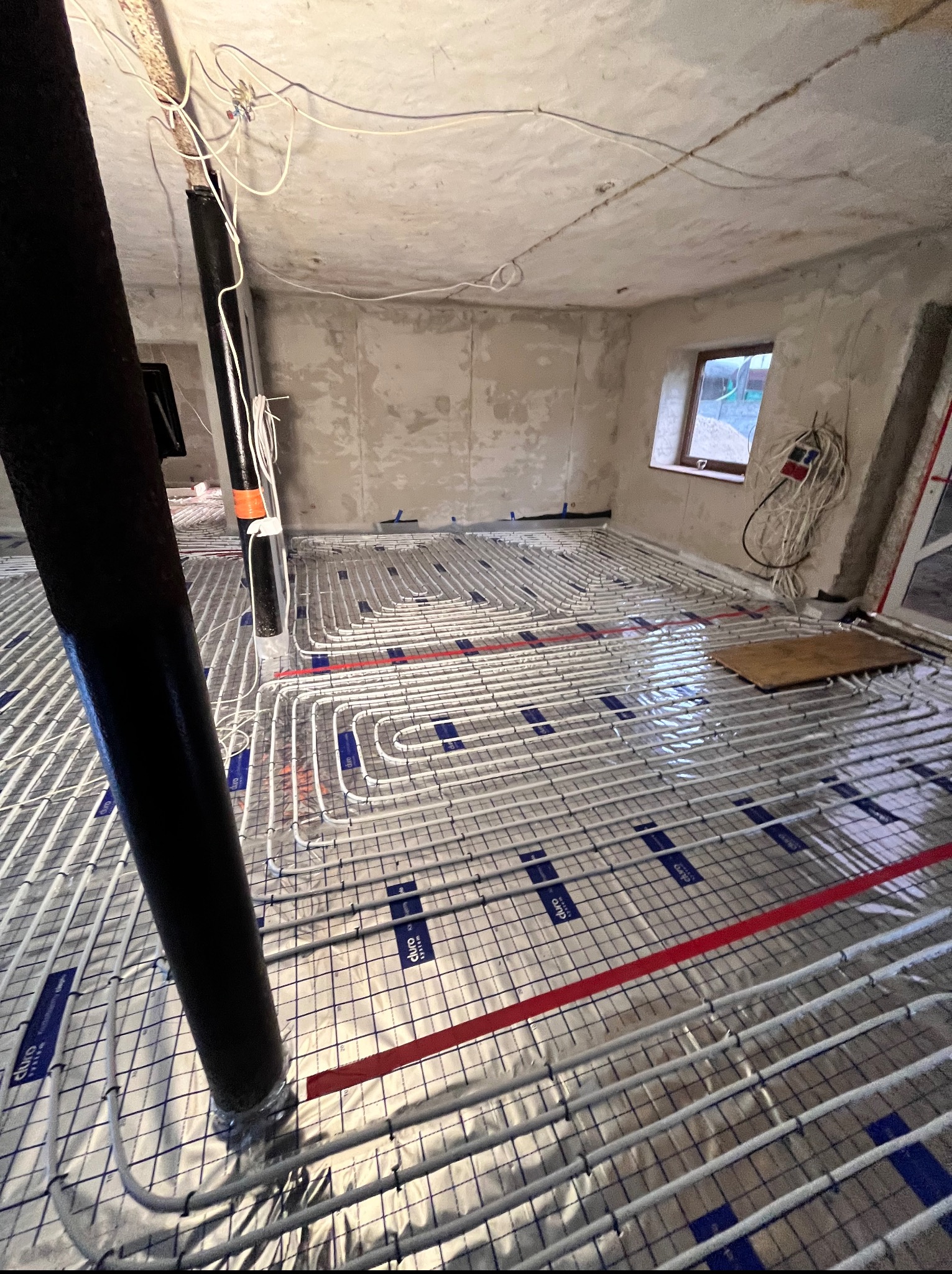 Instalacja ogrzewania podłogowego w surowym wnętrzu. Białe rurki ułożone na srebrnej folii, widoczne przewody elektryczne i okno. Etap prac wykończeniowych.